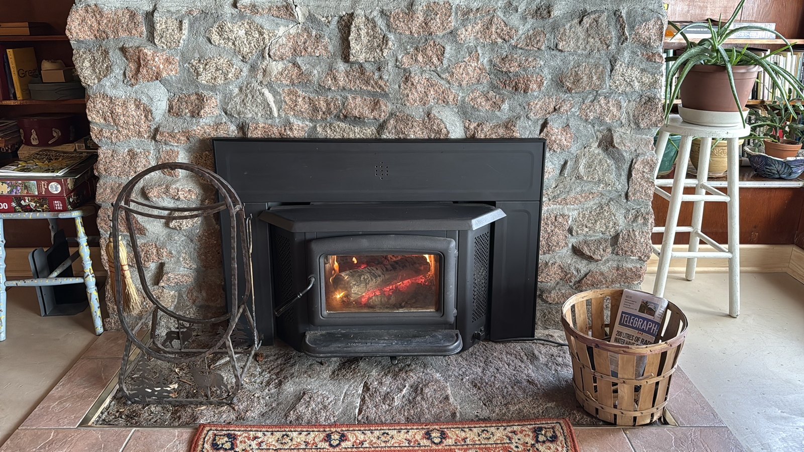 A fire burning in a wood stove insert set into a stone fireplace, with a firewood rack on one side of the hearth and a basket of newspaper on the other