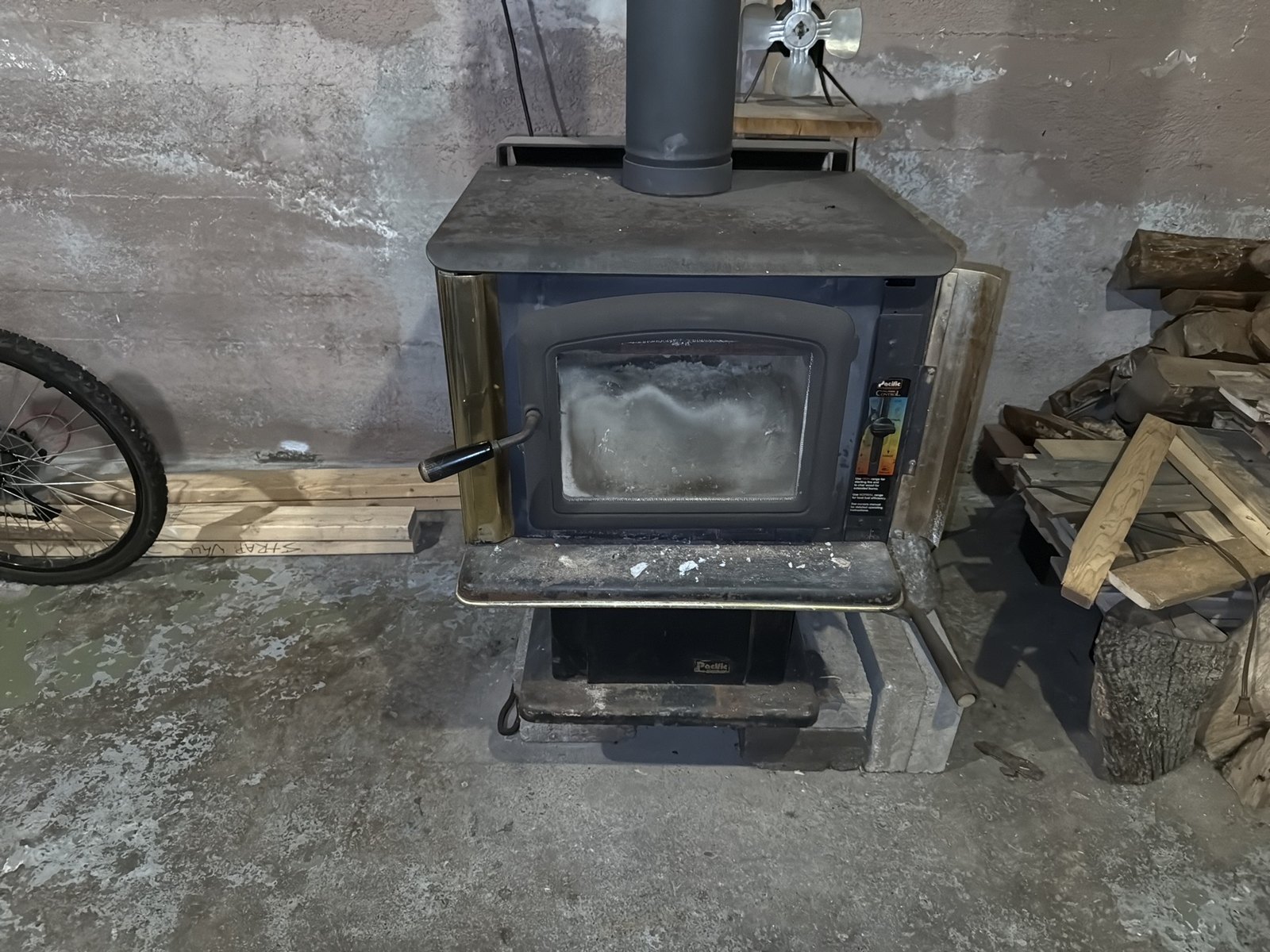 A wood stove in an unfinished basement, standing on concrete against a concrete wall, with its flue pipe rising from the top and firewood stacked beside it