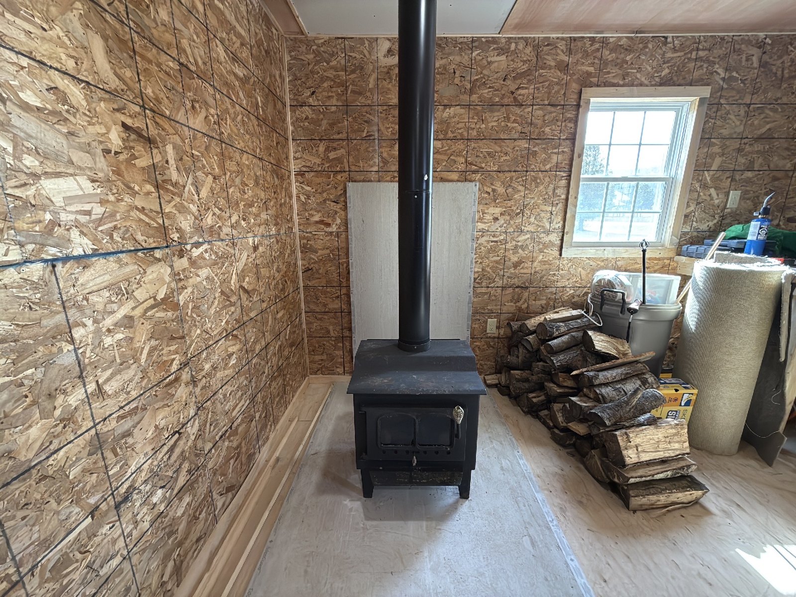 A wood stove in a garage with unfinished walls, its black flue pipe running straight up through the ceiling, with firewood stacked to one side