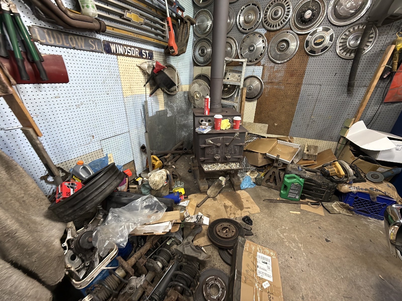 A wood stove in a working garage, with car parts, tools and fluid containers on the floor around it and hubcaps on the pegboard wall behind